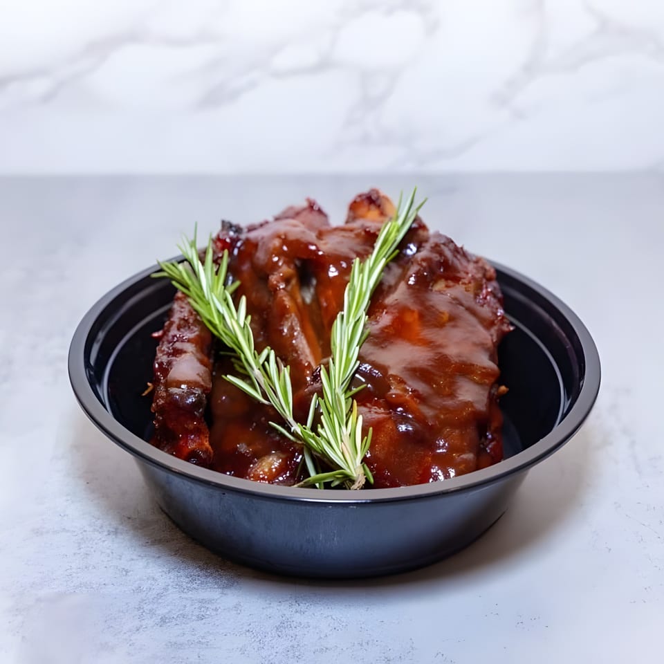 Haitian BBQ ribs with a rich sauce and rosemary sprigs in a black bowl.